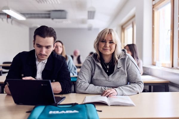 Tva elever sitter vid sina bankar i klassrummet och en kollar rakt in i kameran och ler.